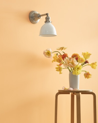 A Sweet Melon-painted wall with single sconce, and a wooden stool with flower-filled vase.
