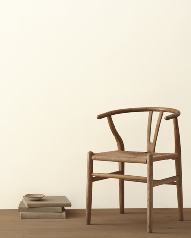 A modern wicker chair and a stack of books topped with a small bowl sit in front of a room painted Mayonnaise.