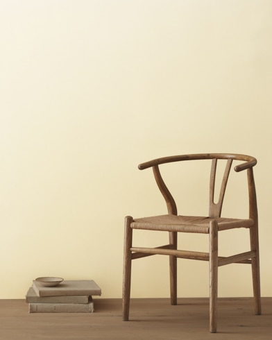 A modern wicker chair and a stack of books topped with a small bowl sit in front of a room painted Woodmont Cream.